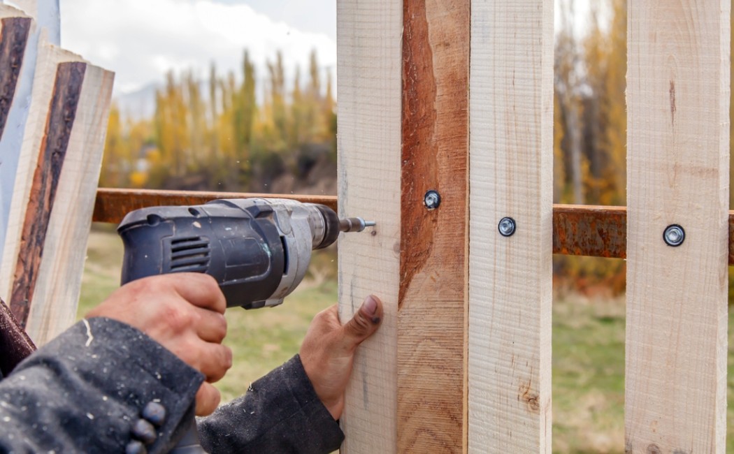 Worker Securing Boards During Fence Installation. Fence Installation in progress as a worker uses a power drill to secure vertical wooden fence boards to a metal support frame outdoors.