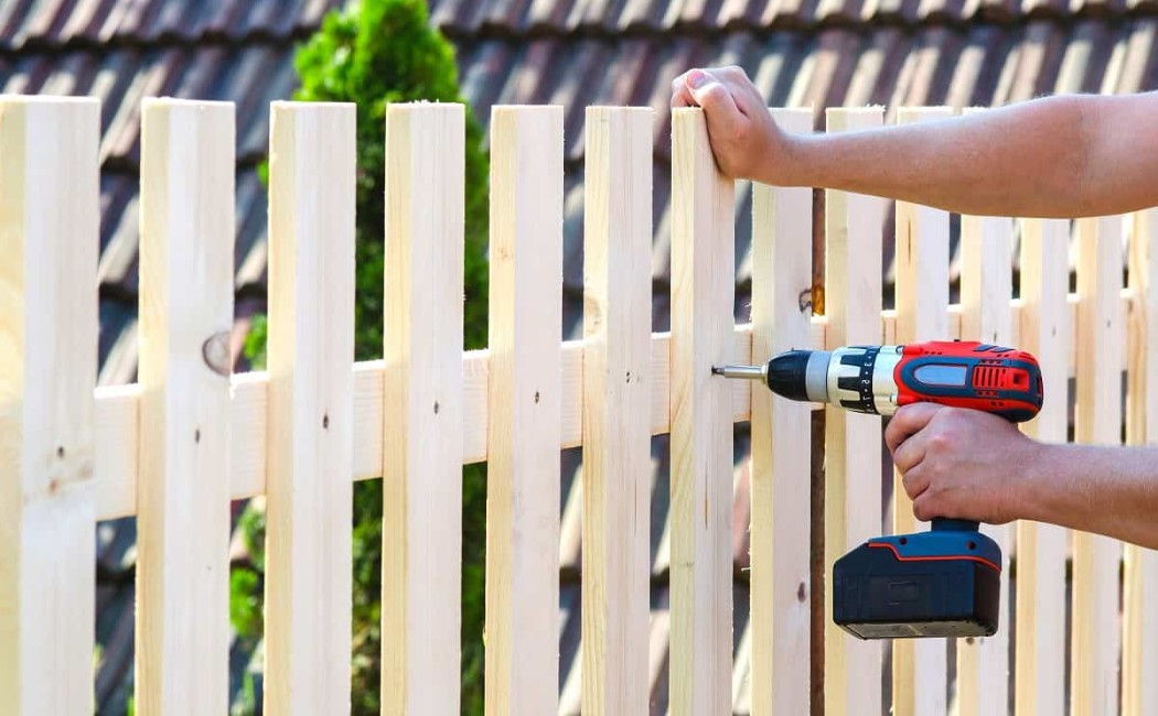 Worker Securing Pickets During Fence Installation. Fence Installation underway as a worker uses a cordless drill to secure wooden pickets on a newly built fence.