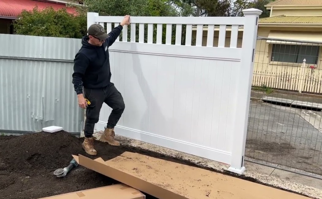 Contractor Inspecting New Vinyl Fence Installation. Fence installation service in progress as a contractor inspects and adjusts a newly installed white vinyl fence panel in a residential yard.