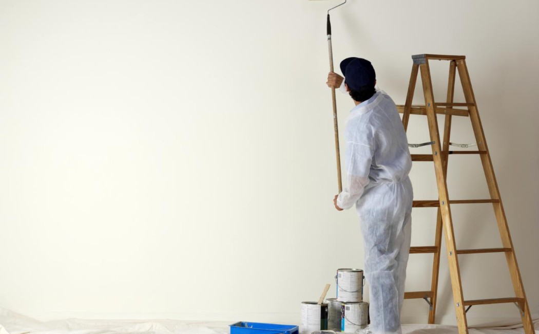 Interior painter applying fresh paint to a large interior wall with a roller while standing beside a wooden ladder in a prepared room.