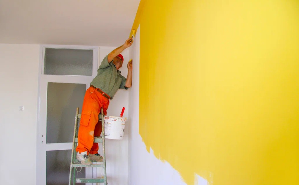 Interior Painting Contractor on Ladder Inside Home. Skilled interior painting contractor applying bright yellow paint to an interior wall using a roller while standing on a ladder.